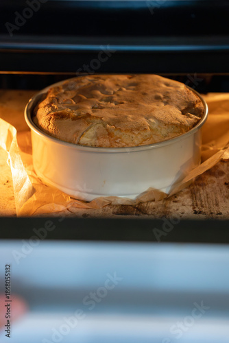 Apple charlotte cake rises in a round baking pan inside the oven, showing golden crust, parchment edges, and warm baking light