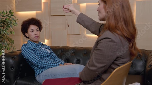Mature woman psychologist hypnotizing pretty African American female patient using pendulum, to create state of focused attention and increased suggestibility in treatment of psychological disorder.
