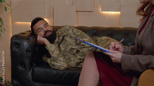Portrait of relaxed African American male army soldier in military uniform laying on sofa, talking with female counselor under hypnosis, sharing positive reminiscences during psychotherapy session.