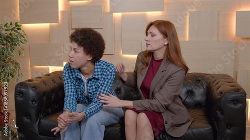 Caring mature woman mental health professional sitting on sofa, consoling depressed African American female patient, expressing understanding, care and support during psychotherapy session.