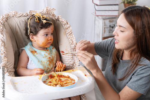 Funny messy baby girl eating spaghetti with tomato sauce on high chair while mother trying to feed at home. Cute toddler kid with dirty face enjoying lunch lifestyle concept.