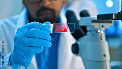Foto A scientist in a lab coat and blue gloves examines a red sample under a microscope, highlighting precision in a clinical research setting