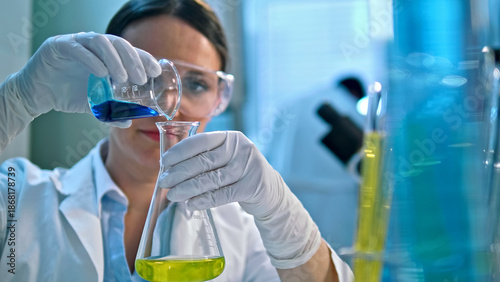 Schilderij op canvas A scientist in a lab coat and gloves carefully pours a blue liquid from a beaker into a flask containing a yellow substance, in a well-lit lab with various chemical containers