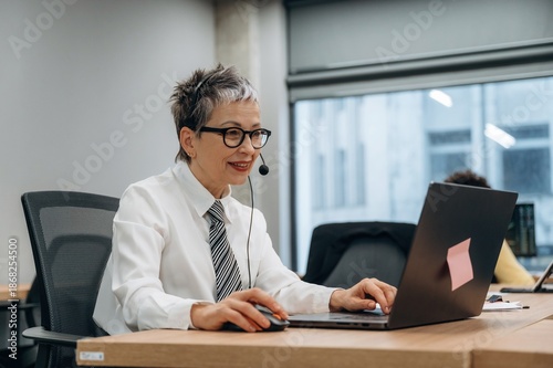 Senior businesswoman with headset working on laptop in modern office environment, showcasing professionalism and dedication to remote communication