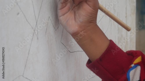 a small boy's hand is drawing or scribbling on a white wall using a simple wooden pencil
