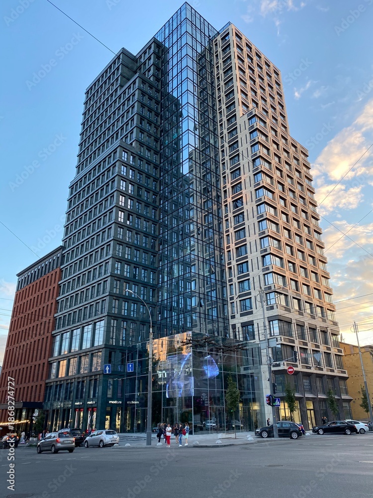 Obraz premium Modern high-rise residential building in an urban city environment during daylight. Contemporary architecture with glass facade and geometric design, surrounded by streets and pedestrians.