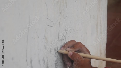 a small boy's hand is drawing or scribbling on a white wall using a simple wooden pencil