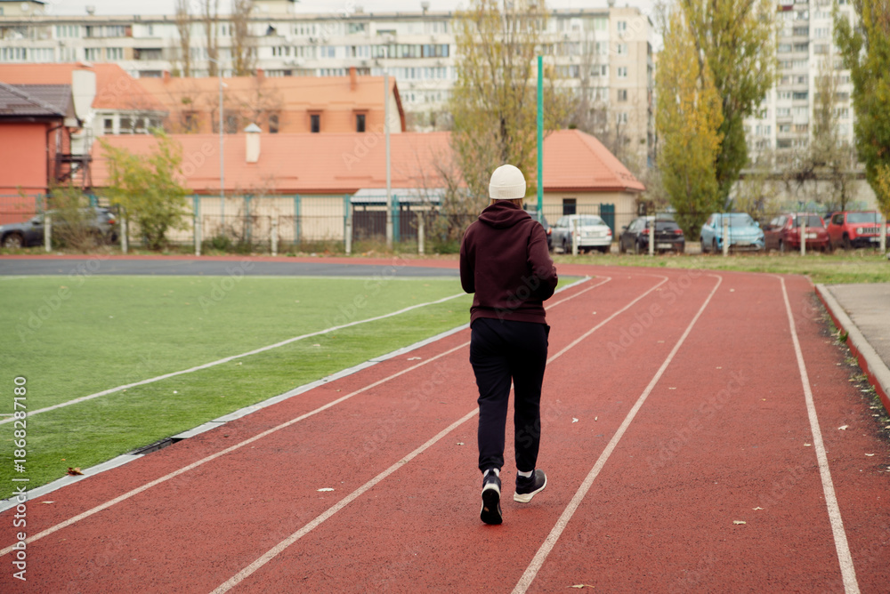 Fototapeta premium Rear view of a person in athletic wear walking on a red running track at an outdoor stadium during autumn. Focus on health, exercise, and urban fitness activity.