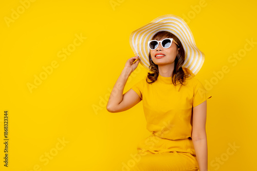 Beautiful happy young Asian woman in vibrant yellow outfit wearing a wide-brimmed straw hat and white sunglasses, smiling cheerfully against a solid yellow background, summer vacation
