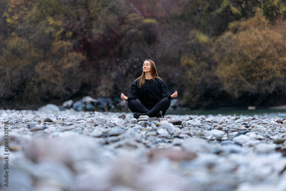 Fototapeta premium Young woman practicing meditation on rocky riverbank, surrounded by nature, with flowing water and autumn foliage, embodying tranquility and mindfulness in a serene environment