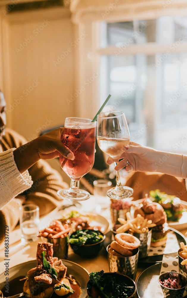 Naklejka premium A group of people are celebrating with drinks and food. They are holding up their glasses and toasting to each other. The table is filled with various dishes, including a plate of fries and a sandwich