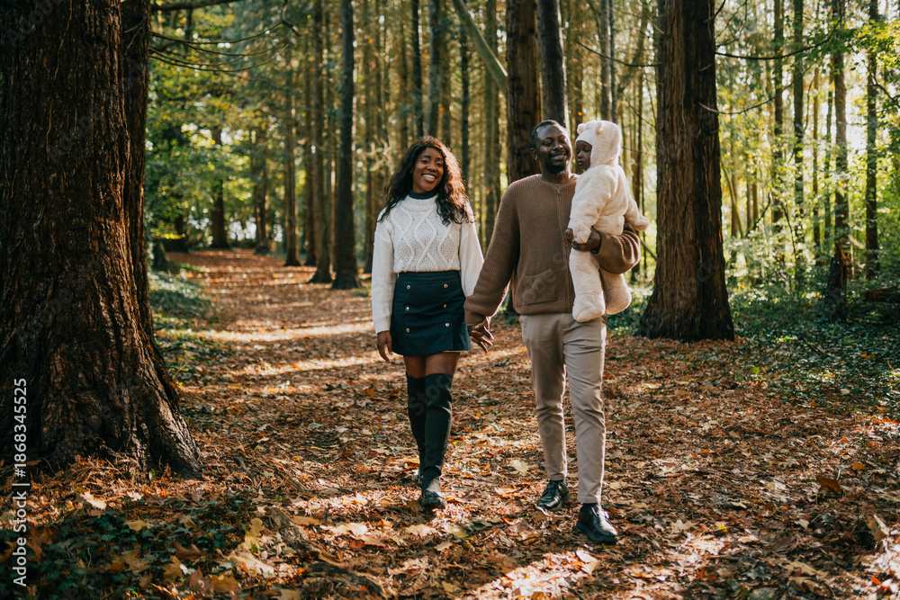 Fototapeta premium A couple holding a baby in a forest. The baby is wearing a white jacket. The couple is walking on a path in the woods