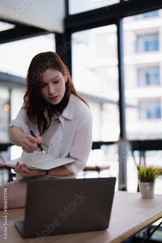 Free Download Young woman collaborating remotely, reviewing notes in her notebook while looking at a laptop, embodying productivity and focus in a bright, contemporary workspace - GFXLoad