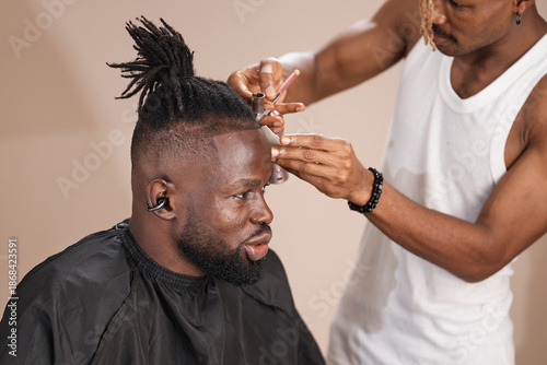 Fotografía Close-up studio shot of a professional barber using an airbrush tool and guide card to shape the hairline of an adult African man with dreadlocks and a beard wearing a black barber cape