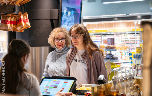 Two women smiling while ordering drinks and interacting with a cashier at a retail counter inside a brightly lit modern airport store