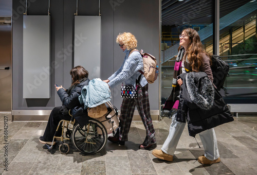 Tablou pe pânză Three women walking through the airport, one using a wheelchair and being assist