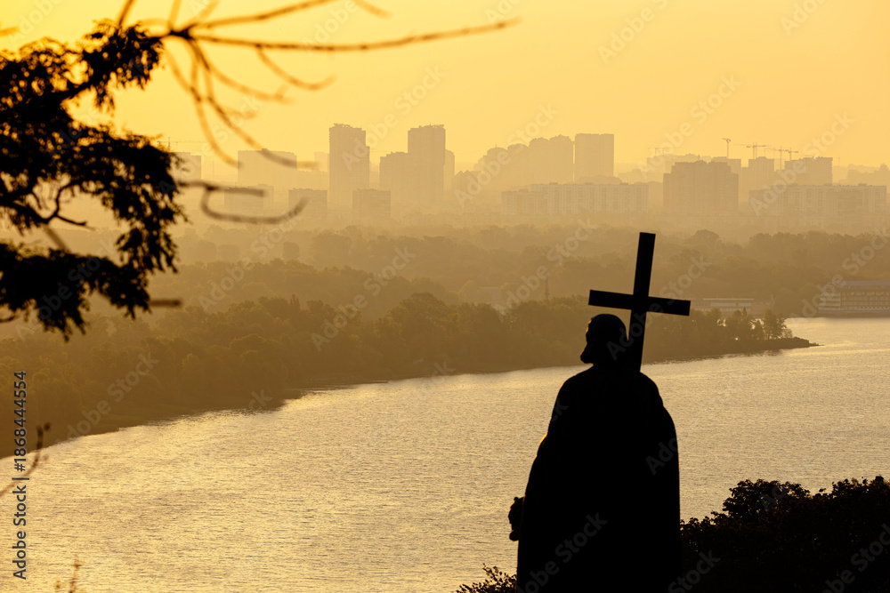 Fototapeta premium Saint Volodymyr Monument silhouette with cross overlooking Dnipro river at sunrise. Hazy city panorama at dawn with residential buildings and forest island under warm morning light in Kyiv.