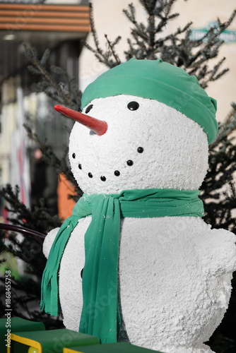 Vertical shot of a Decorative snowman wearing a green hat and scarf with a red carrot nose. Festive Christmas and New Year street decoration with blurred urban background.