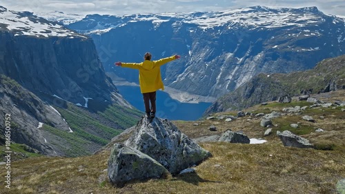 Woman Hiker standing on rock overlooking fjord at Trolltunga in Norway. Person in yellow jacket enjoying dramatic mountain cliffs and deep blue water during a summer hiking adventure.