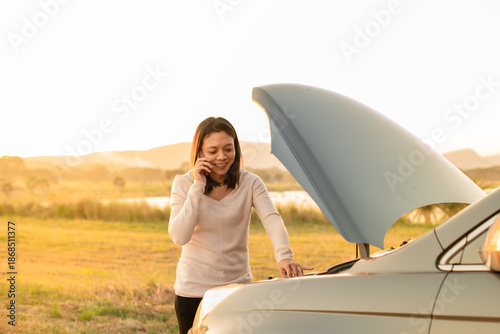 Photos A woman smiling and looking relieved while talking on her mobile phone next to her car with the hood open, suggesting a successful call for help or fixing a minor engine problem