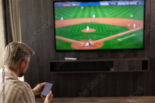 Photography Caucasian man holding smartphone with sports bets app while watching baseball ma