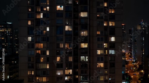 Night view of urban apartment building with lights on in different windows and city lights shining in the background during nighttime in a metropolitan area