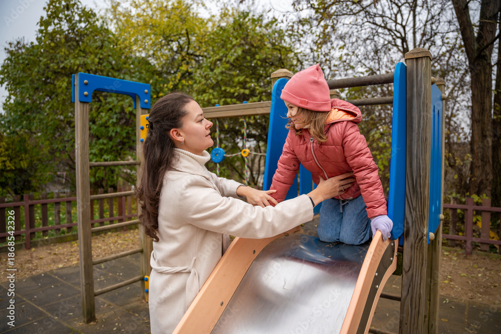 Obraz premium Mother Guiding Daughter Down Playground Slide Careful Hands On Jacket, Shiny Metal Slide, Autumn Leaves, Supportive Posture, Smiling Trustful Moment
