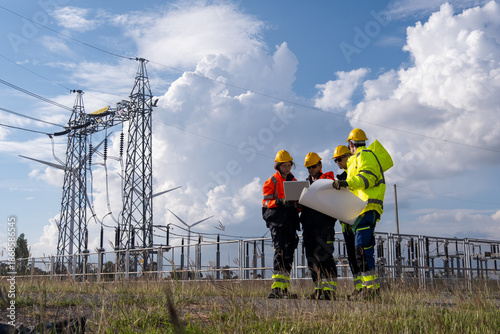 Workers discuss plans at a power station with transmission lines in the background during the day
