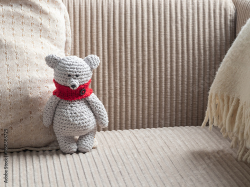 Φωτογραφία Handmade grey crochet teddy bear with red scarf sitting on a beige corduroy sofa, next to a cream pillow and white blanket