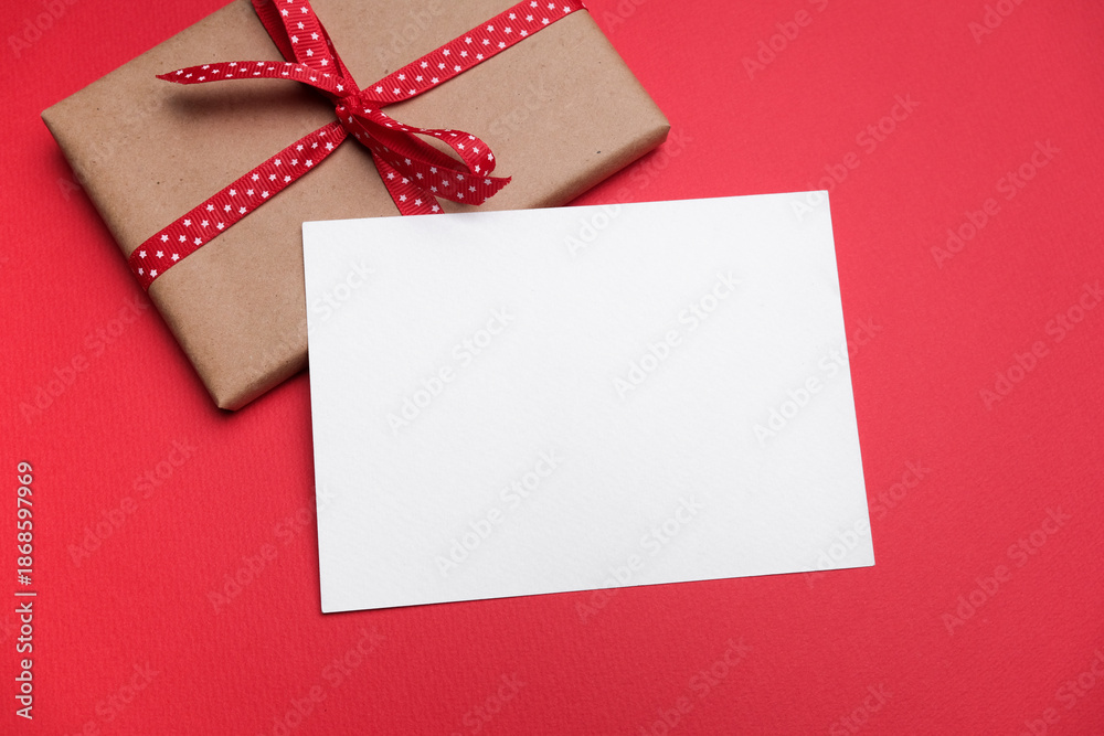 Fototapeta premium Blank paper greeting or invitation card mockup with copy space with gift box featuring a red ribbon on a vibrant red background for festive occasions