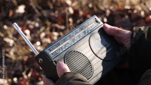 Close-up of male hands holding an old black portable radio receiver with extended antenna. A man tuning a vintage device outdoors against a blurred background of autumn nature.

