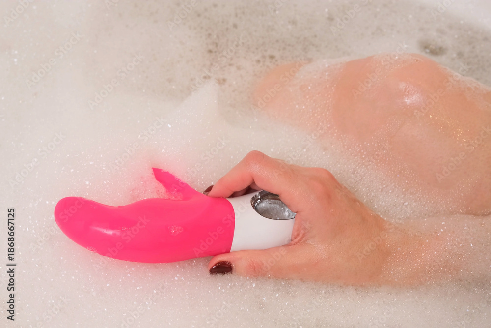 Fototapeta premium Female hand lowers pink sex toy onto soap suds filling tub