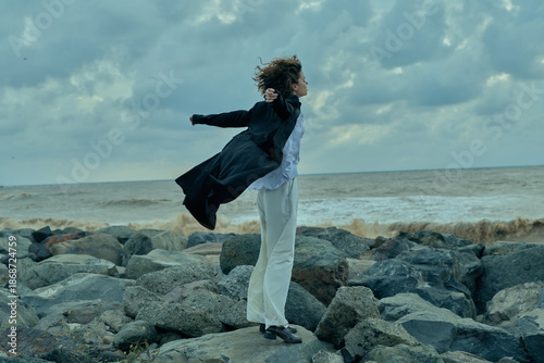 Fotografi Woman balancing on a rocky shoreline wearing a flowing coat, wind whipping hair,