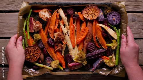 Top view of hands placing a tray of roasted vegetables like beets, carrots, and broccoli on the table. Healthy food for a family Sunday roast dinner