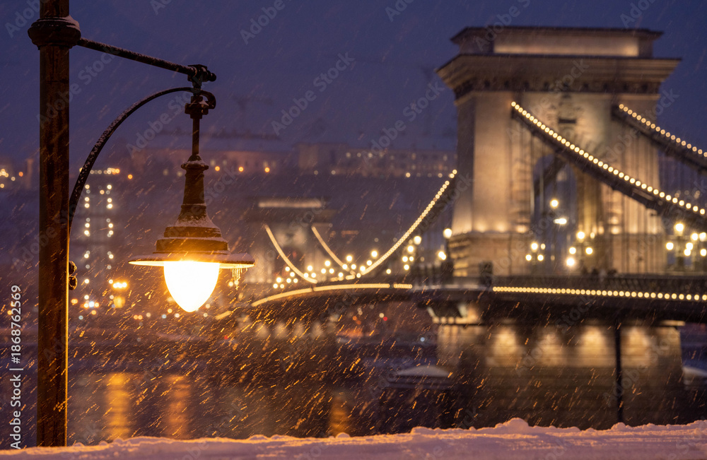 Fototapeta premium Budapest, Hungary - January 6, 2026: The historic Szechenyi Chain Bridge glows with golden lights during a heavy evening snowfall in Budapest, framed by a vintage street lamp and snow-covered