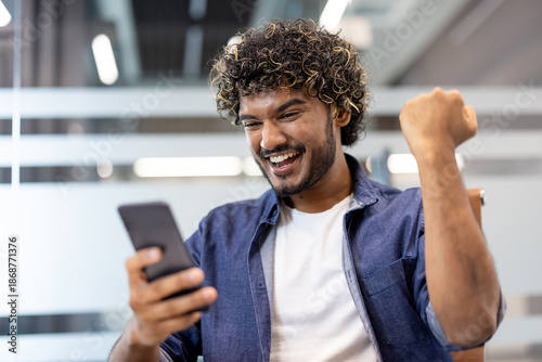 Close-up photo of a happy young Indian man in the office, looking at his phone screen and rejoicing in victory