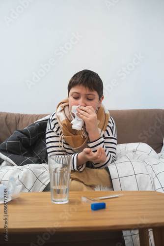 Sick child sitting on sofa wrapped in blanket, holding tissue to nose, winter illness concept, home interior, cold or flu symptoms.