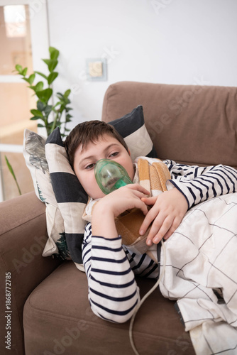 Boy wrapped in blanket using inhaler mask, sitting on sofa at home, recovering from illness, glass of water nearby, cozy indoor setting.
