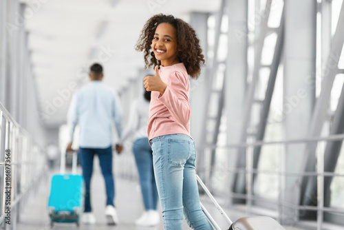 Travel Concept. Portrait of smiling excited African American teenage girl walking with suitcase at airport terminal corridor, showing thumbs up sign gesture, looking back at camera, enjoying vacation