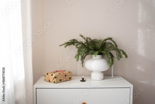 inimalist winter decor: white textured vase with lush fir branches, gift box wrapped in festive paper with red ribbon, small candle on white dresser near sheer curtains. Cozy indoor scene.