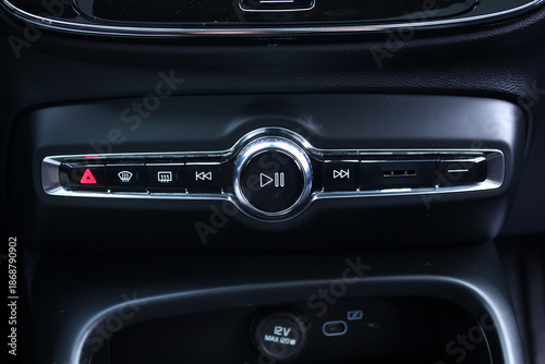 Close-up of a modern car's center console with media and function controls. The black panel features a central play and pause knob, hazard and defrost buttons, all highlighted by chrome accents.