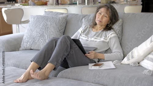 Thoughtful woman sitting on living room sofa relaxing looking out window thinking working at home laptop on lap in peaceful home