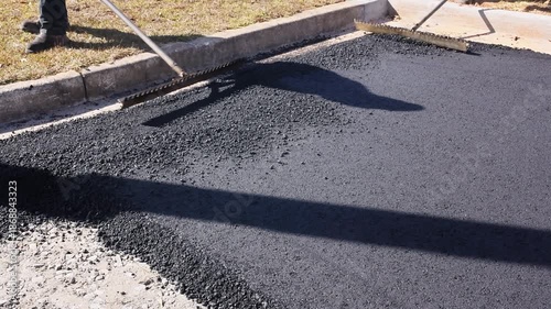 Workers use tools to spread hot asphalt on ground for road repair at construction site