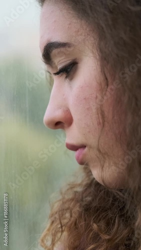 Close-up profile of a pensive young woman with acne looking out a window, feeling sad and lonely. Vertical video.