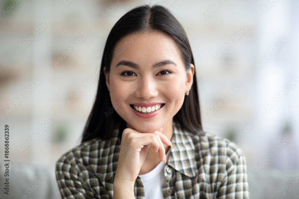 Fototapeta premium Closeup potrait of attractive long-haired asian woman in cozy outfit chilling at home, smiling at camera, copy space. Home interior, millennials lifestyle, relaxation and leisure concept