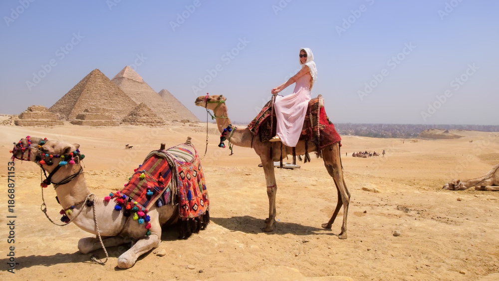 Fototapeta premium A joyful traveler rides a camel in the Egyptian desert with the iconic Giza pyramids in the background, capturing the perfect vacation moment.