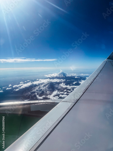 Aerial photograph of the volcanoes, Popocatepetl and Iztaccihuatl covered in snow, seen from an airplane, the wing of the same can be distinguished, with many clouds below the volcanoes.
