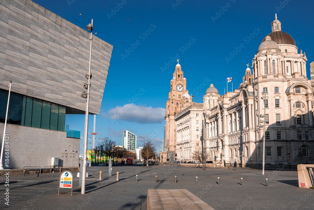 Naklejka premium Winter sun illuminates Liverpool Pier Head. Royal Liver Building and Port of Liverpool Building stand near Museum of Liverpool. Sparse pedestrians cast long shadows.