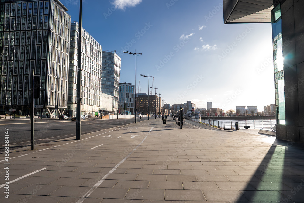 Fototapeta premium Winter afternoon on Liverpool waterfront shows promenade and cycle lane by River Mersey, Pier Head and Mann Island ahead, cranes near Albert Dock, long shadows and reflections.