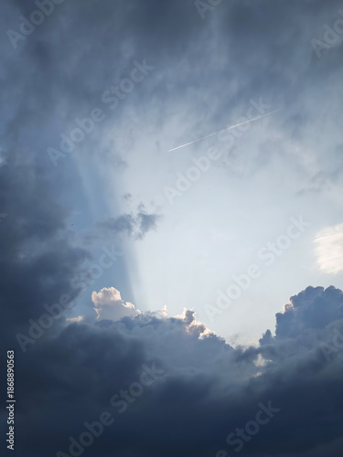 Sun rays breaking through dark storm clouds with a jet plane contrail in the sky.
